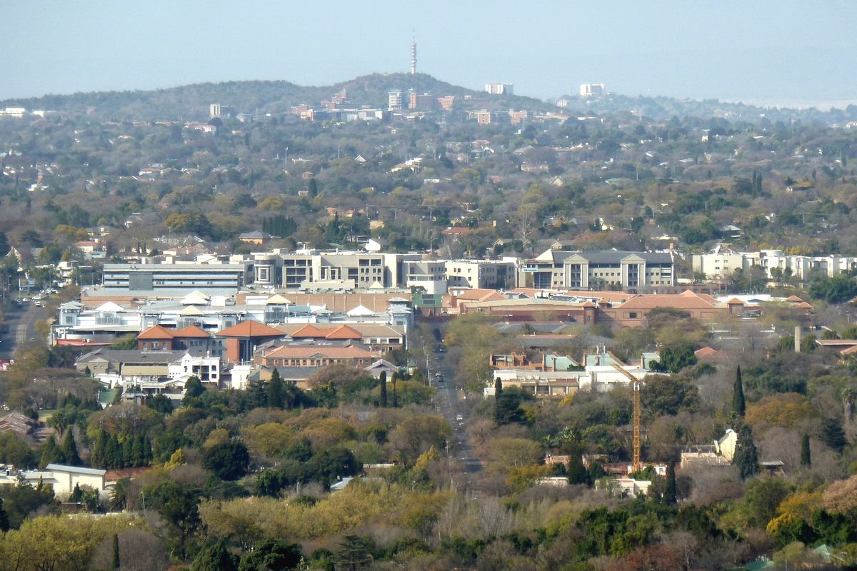 A leafy Pretoria suburb of tree-lined streets and red-roofed buildings, with the Klapperkop tower on the ridge behind.
