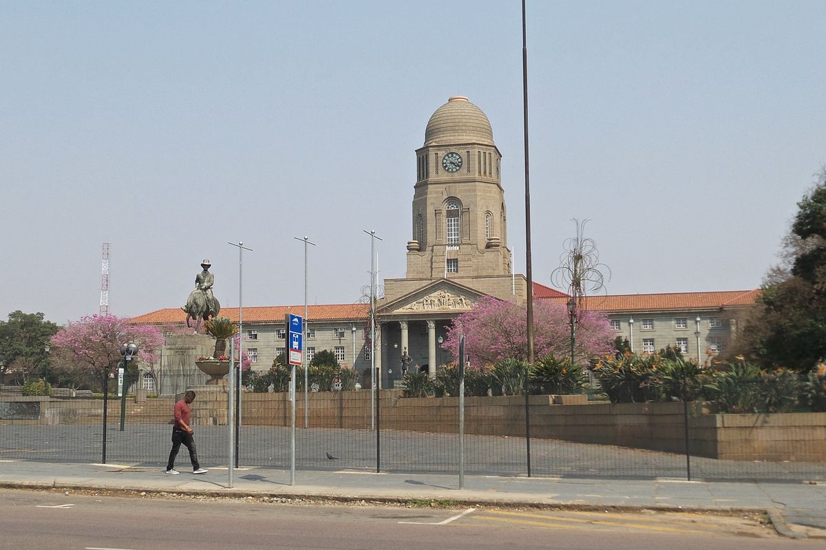 The old Pretoria City Hall: a classical civic building with a clock tower, seen across the street with jacarandas in bloom.